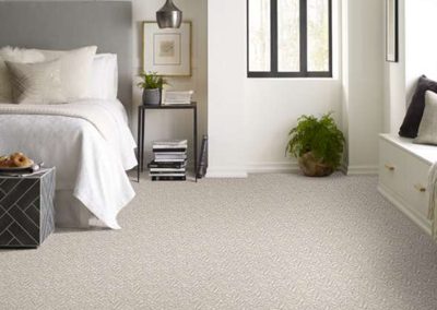 Modern bedroom with a gray bed, white walls, and carpeted floor by Eastman's Carpets Inc. A window and a bench with cushions complete the look, showcasing expert flooring services for a cozy retreat.