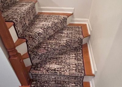 A staircase with a patterned carpet runner on steps and landing, featuring wooden edges and white walls—an elegant flooring solution by Eastman’s Carpets Inc.