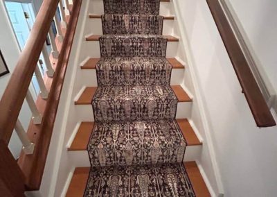 Staircase with a patterned carpet runner by Eastman's Carpets and wooden handrails on both sides in a well-lit hallway, showcasing expert flooring services.