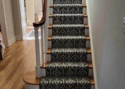 A carpeted staircase with a narrow runner and wooden steps by Eastman's Carpets Inc brightens a hallway with hardwood floors, showcasing expert flooring solutions.