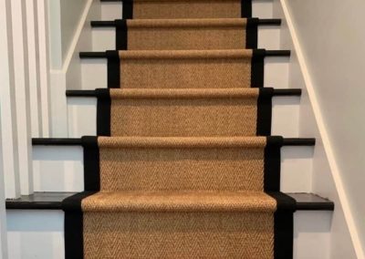 Staircase with a tan carpet runner bordered by black, leading upward between white walls—expertly installed by Eastman’s Carpets Inc, your trusted provider of quality flooring services.