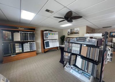 Carpet and flooring samples from Eastman’s Carpets are displayed on racks in a well-lit showroom with beige carpet and white ceiling tiles, showcasing a variety of flooring solutions.